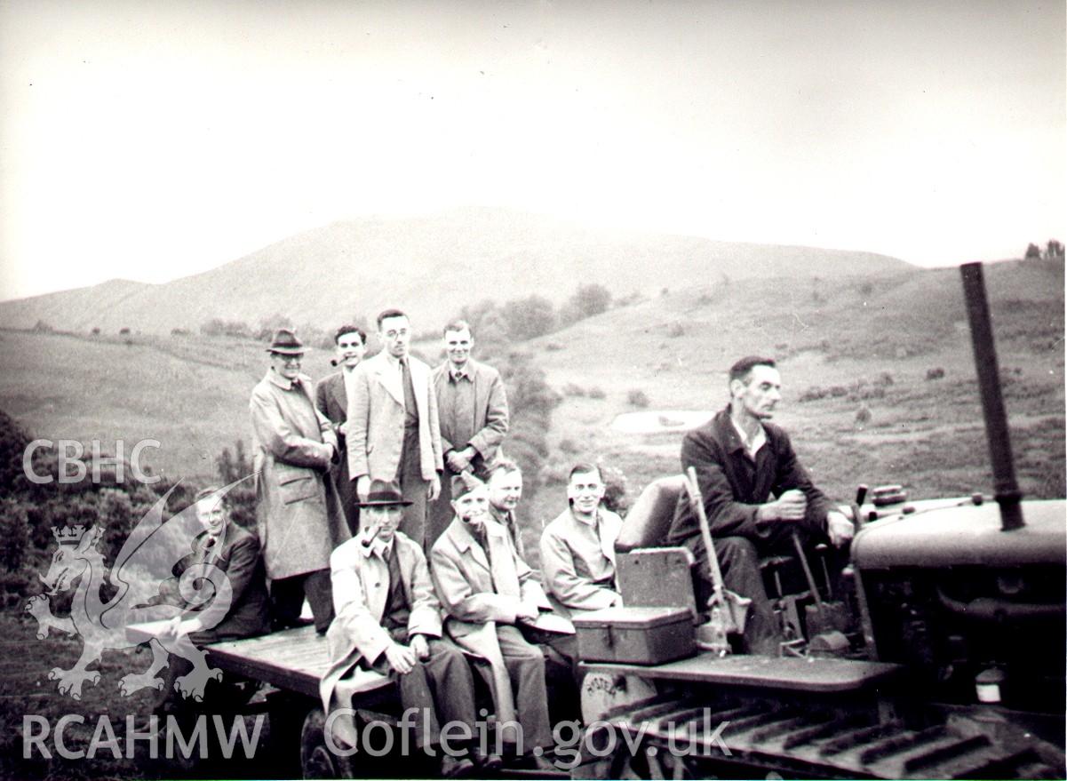 Black and white digital photograph showing group of men on a tractor drawn trailer.