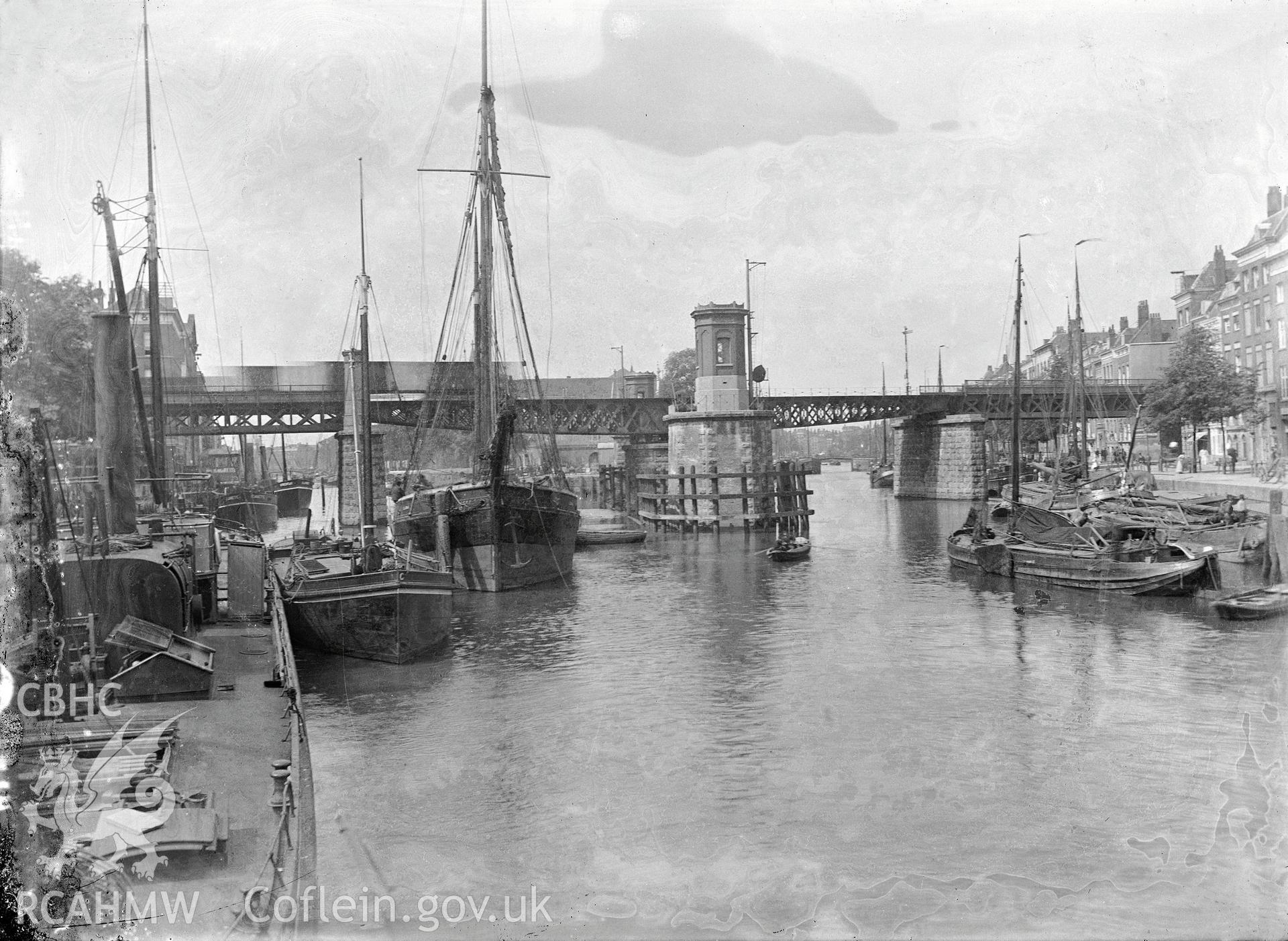 Black and white image dating from c.1910 showing a European harbour or river scene crossed by a bridge, featuring a number of sailing vessels, taken by Emile T. Evans.
