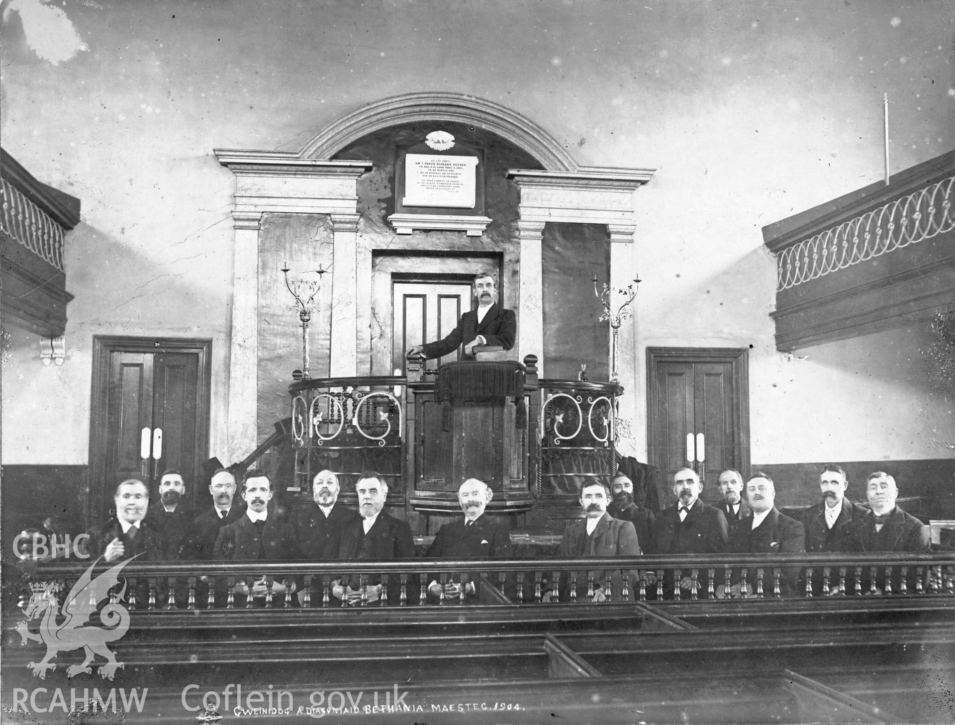 Digitized copy of a black and white photograph showing an interior view of Bethania Chapel, Maesteg, with  the minister and deacons. Part of a collection of material relating to Bethania Chapel, Maesteg, loaned for copying by the Welsh Religious Buildings Trust. The original collection is held by the Glamorgan Record Office.