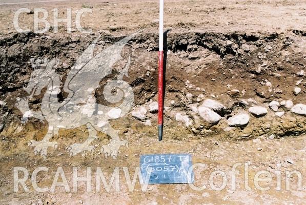 Photograph from excavation of Parc Bryn Cegin, Llandygai, by Gwynedd Archaeological Trust. Detail of machine cut section through (3057), from SSW (numbers refer to context records). Scale 1x1m.