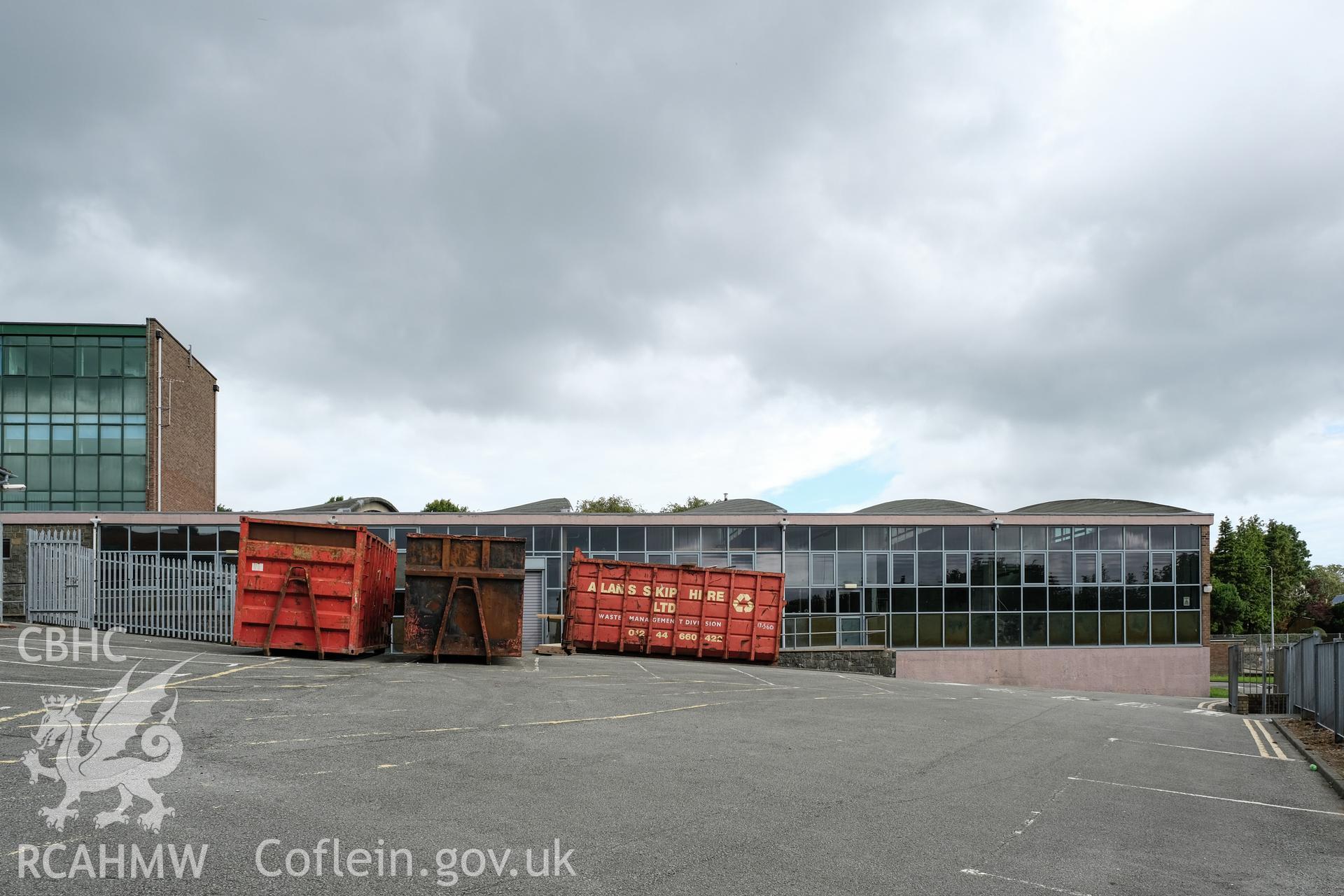 Digital colour photograph of exterior view of a heavily fenestrated wall with skips at Caernarfonshire Technical College, Bangor. Photographed by Dilys Morgan and donated by Wyn Thomas of Grwp Llandrillo-Menai Further Education College, 2019.