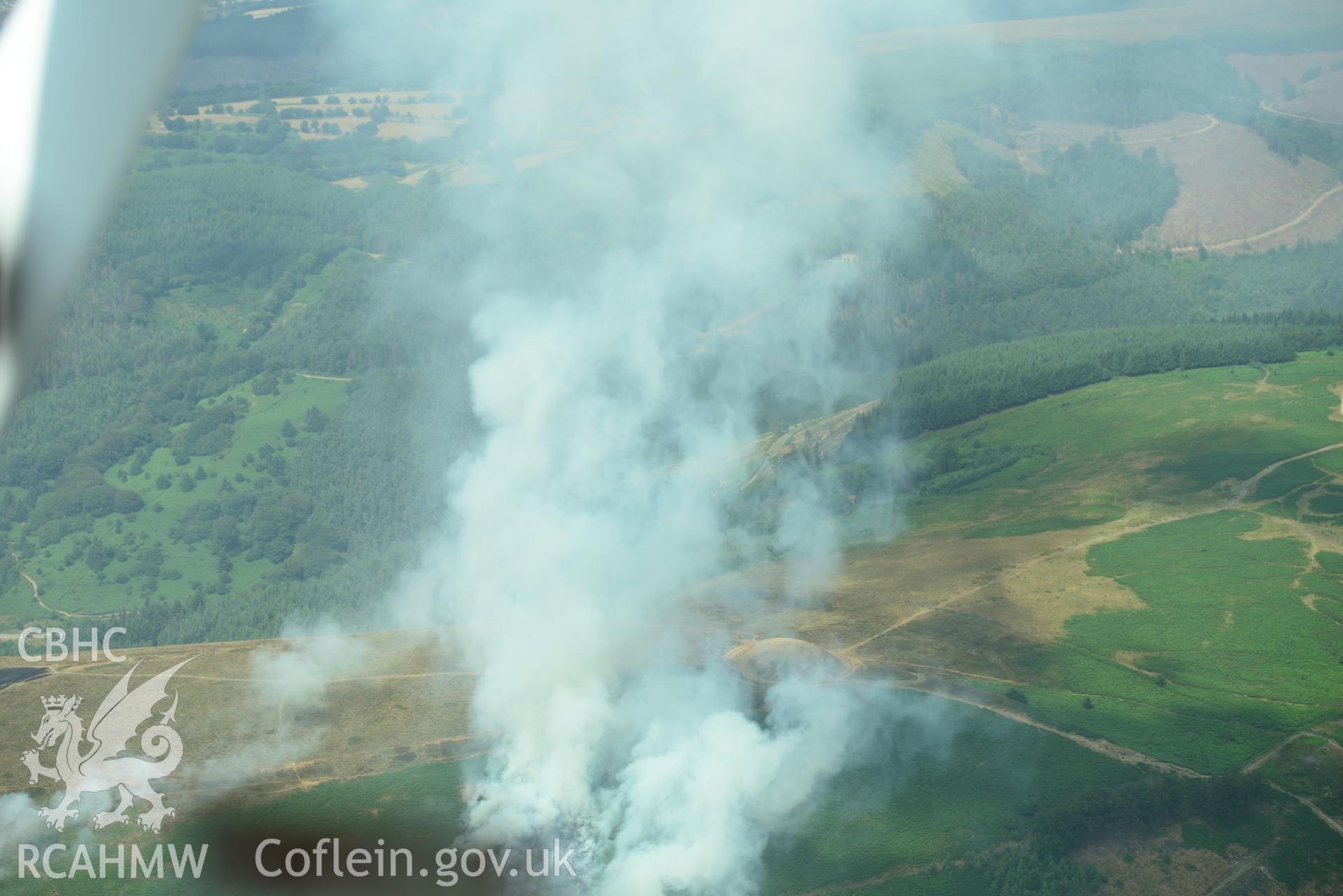 Royal Commission aerial photography of Twmbarlwm motte and bailey during a fire taken on 19th July 2018 during the 2018 drought. The photograph is taken through the window of the aircraft due to the pilot following a flight path to avoid the smoke.