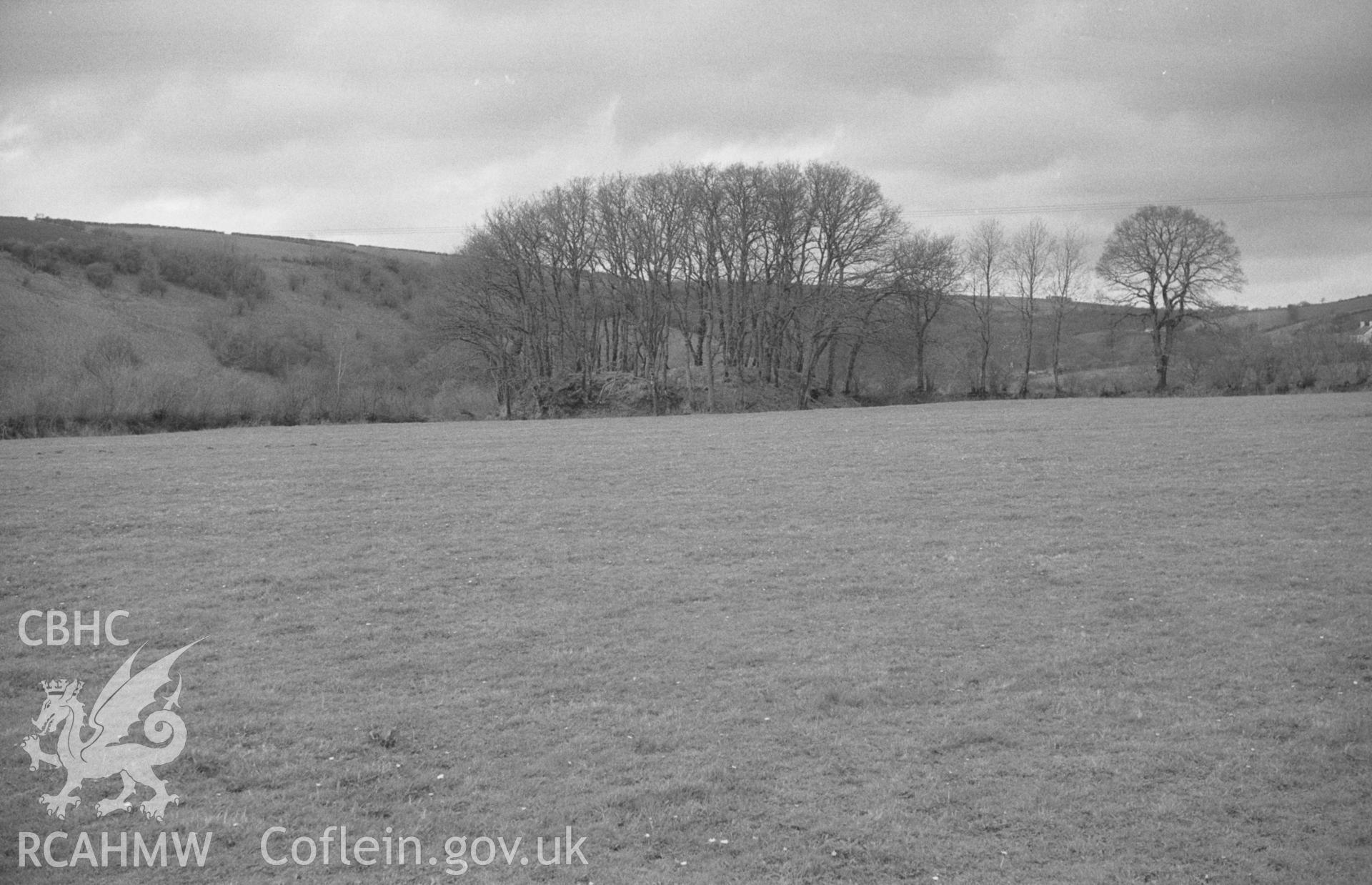 Digital copy of a black and white negative showing Castell Nant-y-Garan motted, covered in trees, seen from near the road in Llandyfriog. Photographed by Arthur O. Chater in April 1966 from Grid Reference SN 370 420, looking north north west.