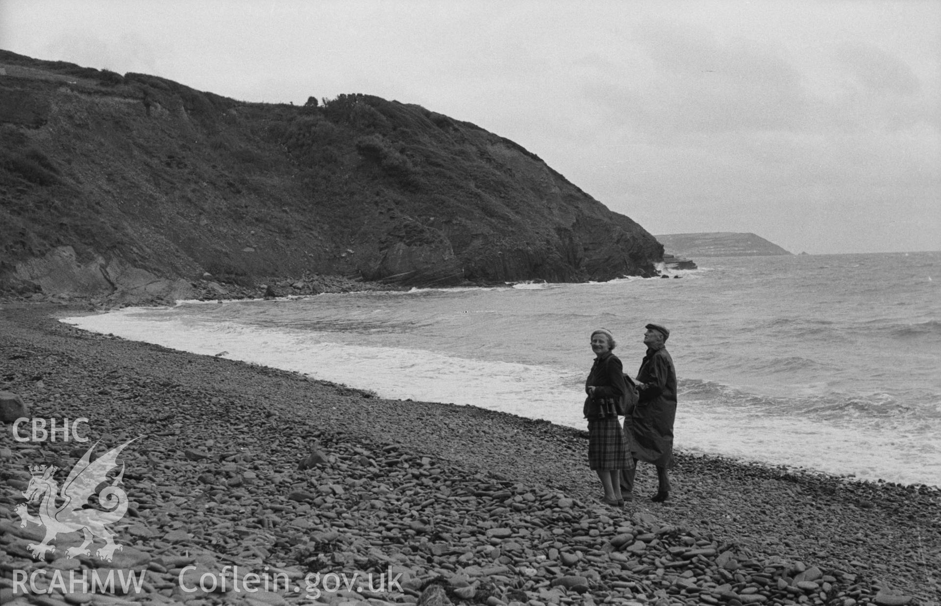 Black and White photograph showing figures at Gilfach yr Halen beach, 200m north east of the mouth of Afon Cwinten, New Quay Head in distance. Photographed by Arthur Chater in August 1962 from Grid Reference SN 437 615 looking south west.