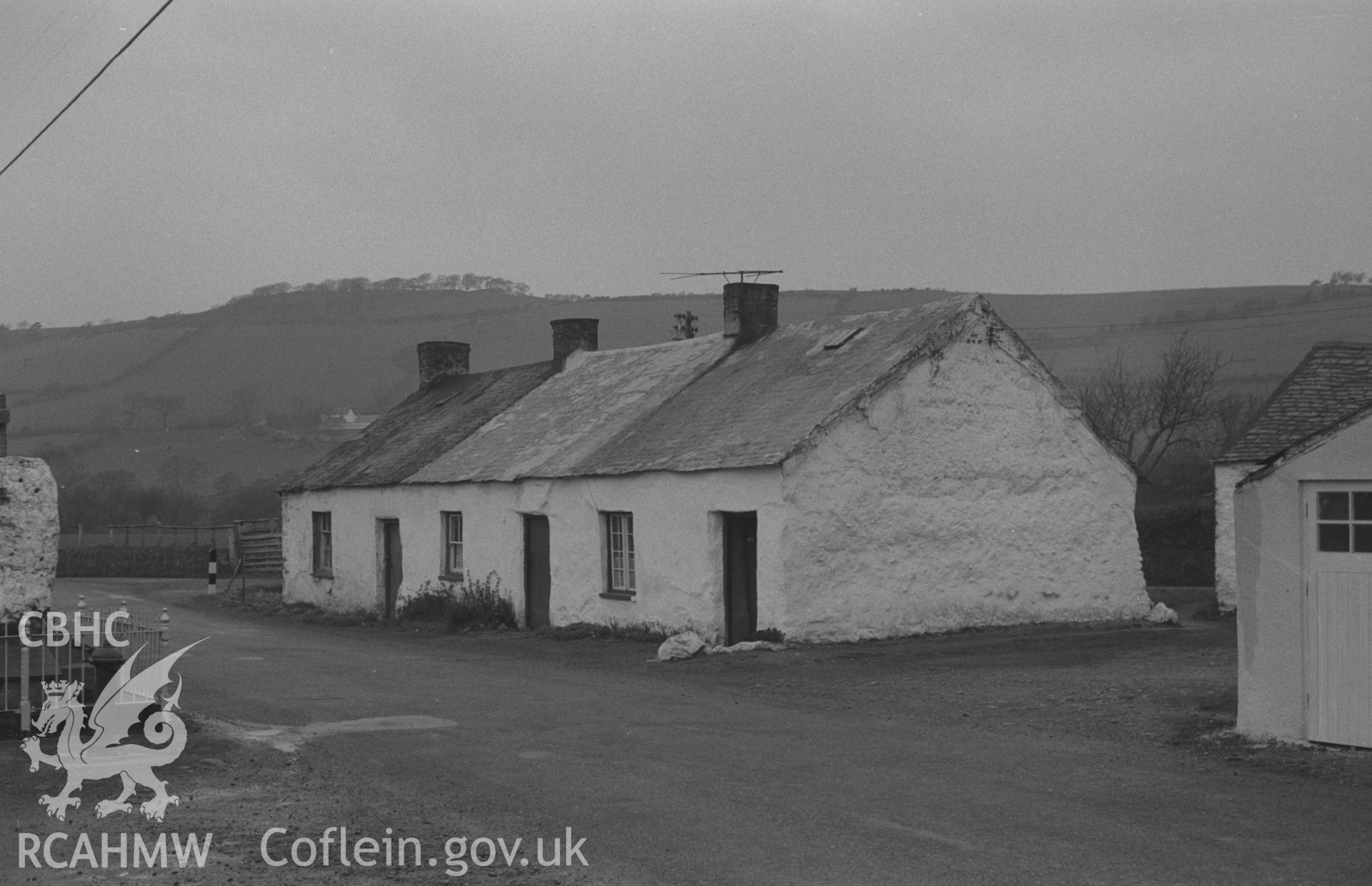 Black and White photograph showing row of mud cottages in the angle of the roads in middle of Penrhyncoch village; Hen Gaer iron-age camp in trees on skyline. Photographed by Arthur Chater, April 1962 at Grid Reference SN642841, looking west north-west.