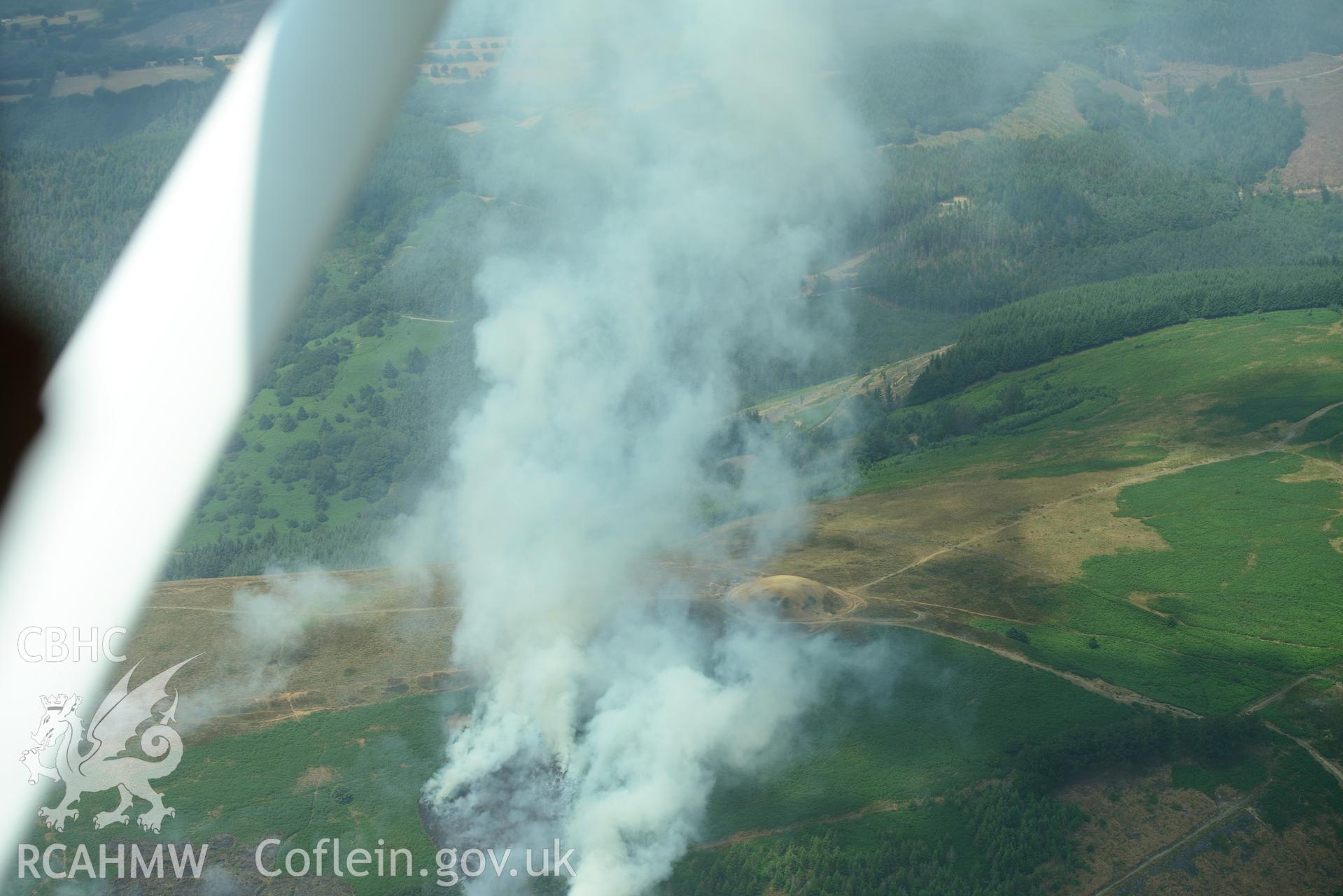 Royal Commission aerial photography of Twmbarlwm motte and bailey during a fire taken on 19th July 2018 during the 2018 drought. The photograph is taken through the window of the aircraft due to the pilot following a flight path to avoid the smoke.