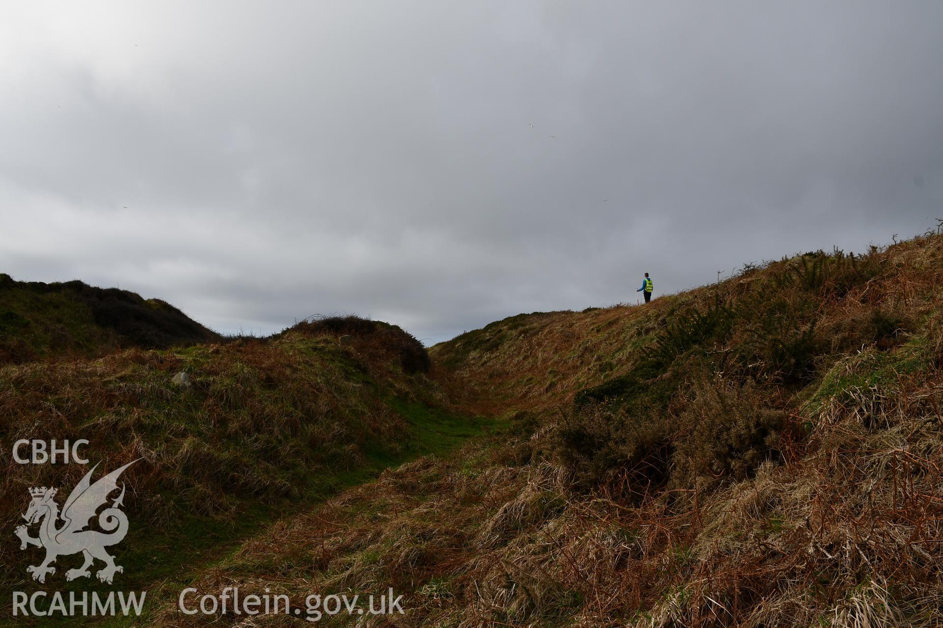 Ramparts and ditches. Person and 1m scale. Camera facing W. Taken by Hannah Genders Boyd. Produced with EU funds through the Ireland Wales Co-operation Programme 2014-2023. All material made freely available through the Open Government Licence.
