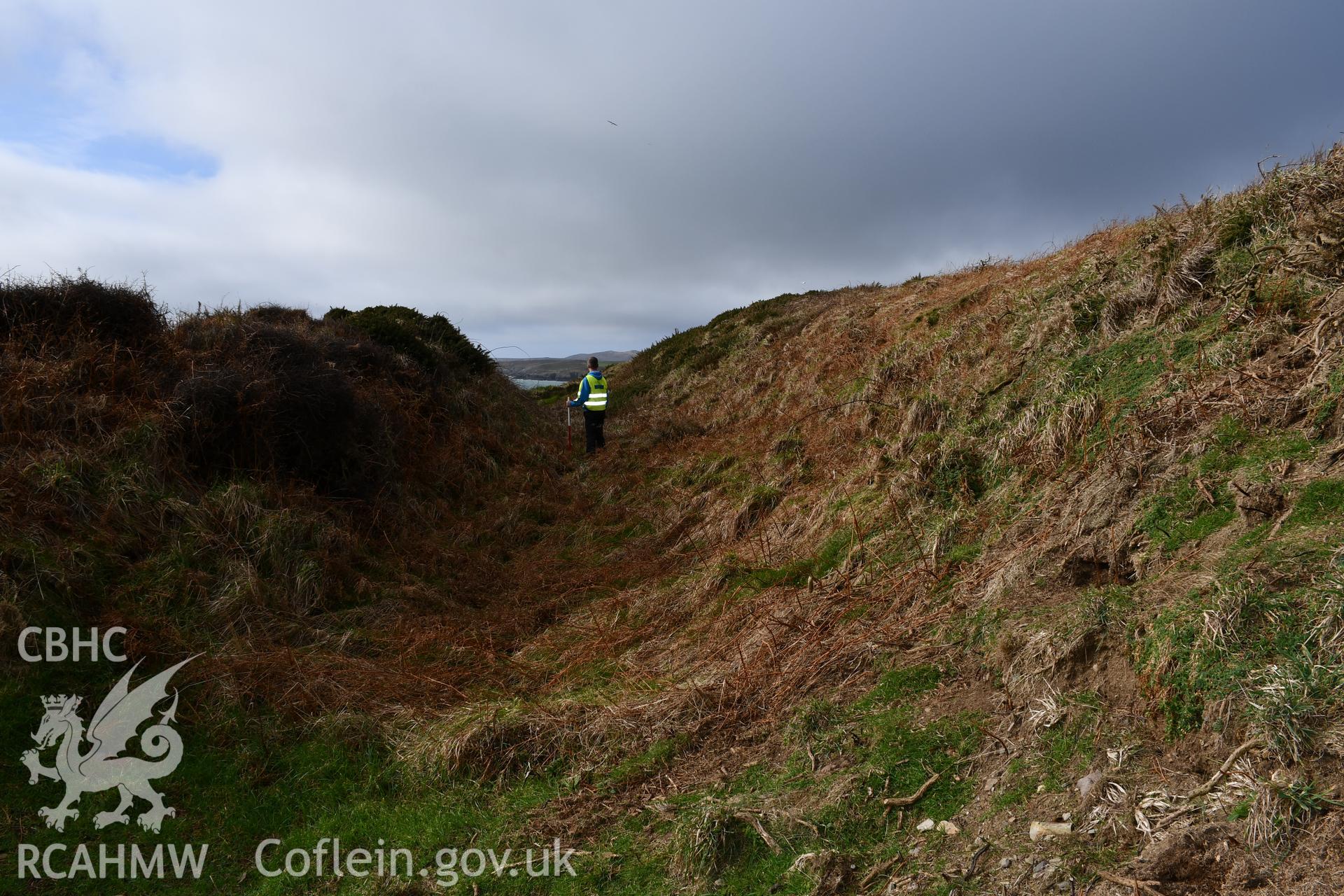 Ramparts and ditch. Person and 1m scale. Camera facing W. Taken by Hannah Genders Boyd. Produced with EU funds through the Ireland Wales Co-operation Programme 2014-2023. All material made freely available through the Open Government Licence.