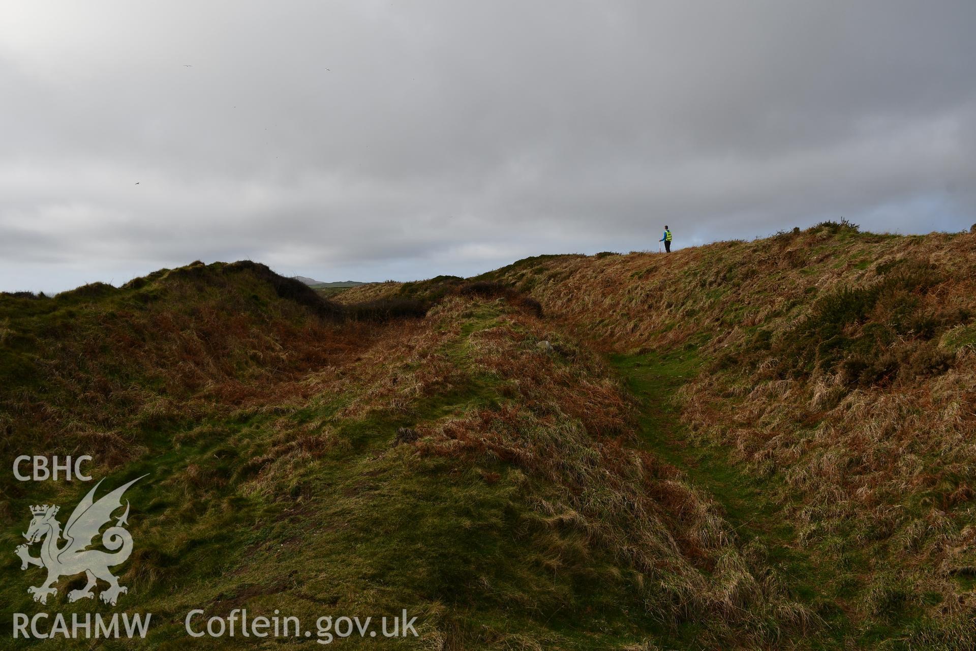 Ramparts and ditches. Person and 1m scale. Camera facing W. Taken by Hannah Genders Boyd. Produced with EU funds through the Ireland Wales Co-operation Programme 2014-2023. All material made freely available through the Open Government Licence.