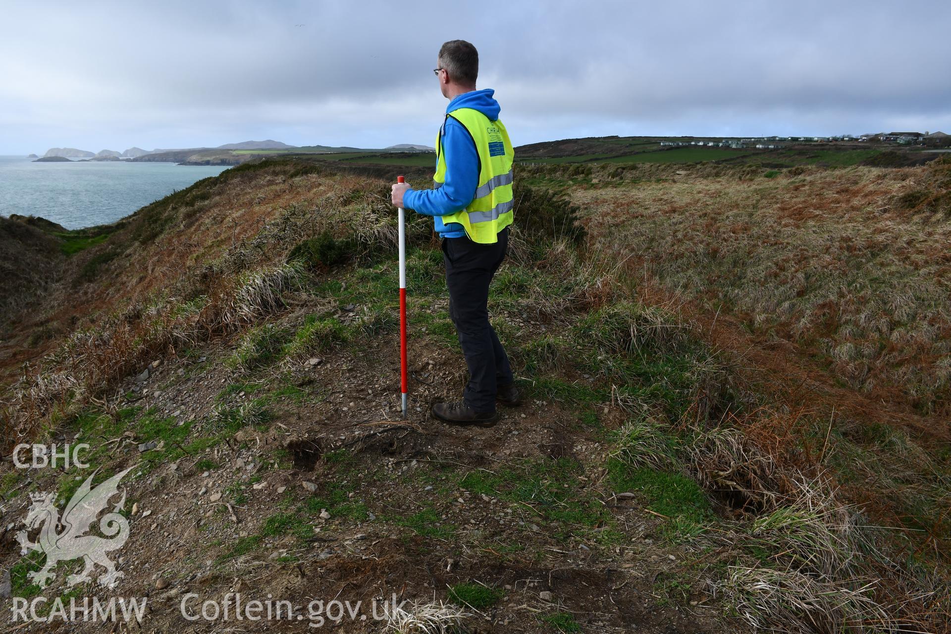 Reinstatement of turf over trench through rampart from Autumn 2021 excavation. Camera facing W. Person and 1m scale. Taken by Hannah Genders Boyd. Produced with EU funds through the Ireland Wales Co-operation Programme 2014-2023. All material made freely