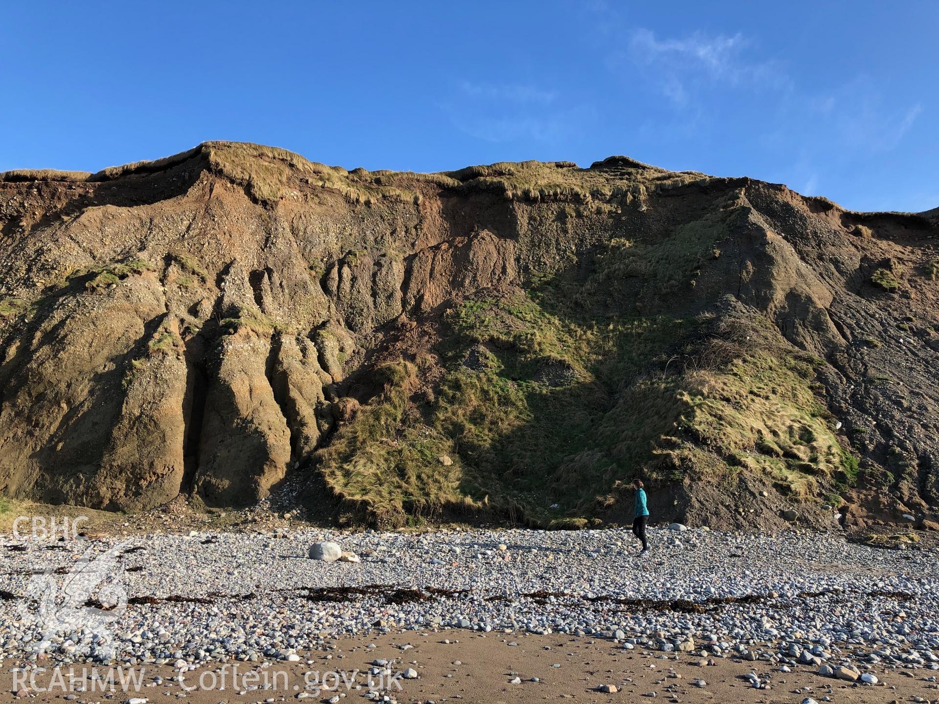 Wide shot of eroding edge showing grooves from water run-off. Figure for scale. Taken by Toby Driver. Produced with EU funds through the Ireland Wales Co-operation Programme 2014-2023. All material made freely available through the Open Government Licence
