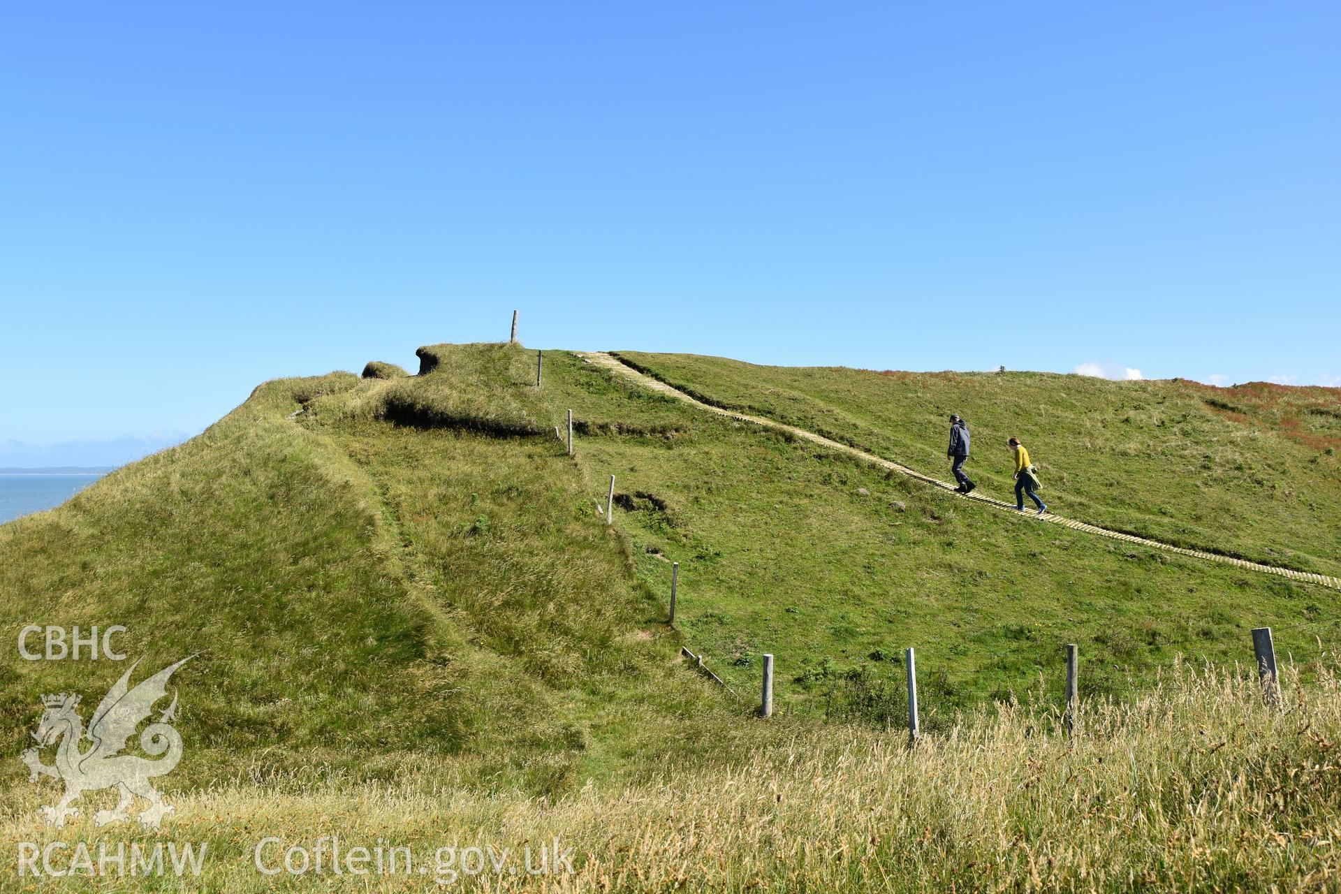 Enclosure and inner ramparts with figures for scale. Taken by Daniel Hunt. Produced with EU funds through the Ireland Wales Co-operation Programme 2014-2023. All material made freely available through the Open Government Licence.