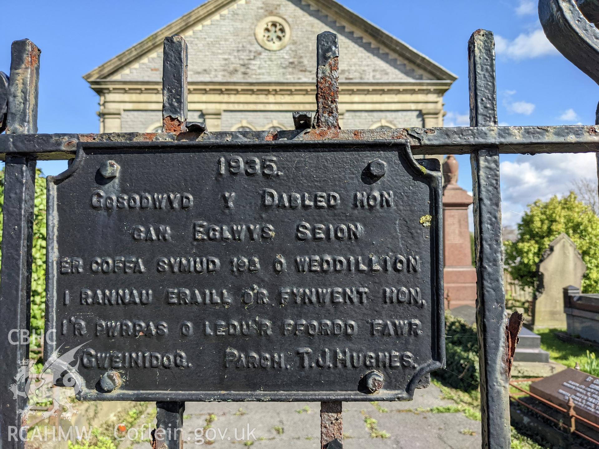Gate sign: '1935. Gosodwyd y dabled hon gan Eglwys Seion er coffa symud 198 o weddillion i rannau eraill o'r fynwent hon, i'r pwrpas o ledu'r ffordd fawr. Gweinidog. Parch. T. J. Hughes'. Translation: '1935. This tablet was placed to commemorate the movin