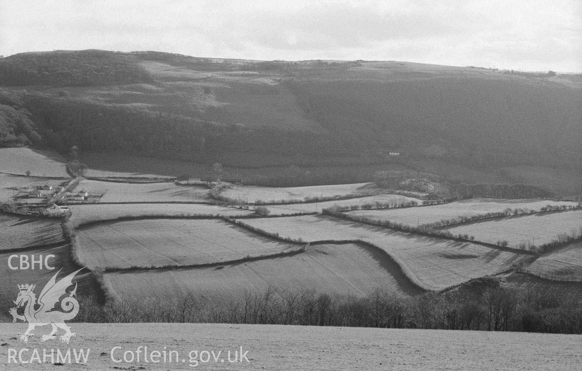 Digital copy of a black and white negative showing view from the lane above Coed Troed-y-Rhiw across the Melindwr valley to Penbryn (just left of centre). Coed Penbryn at top left. Blackhorse House showing white on main road right of centre. Field patterns showing up in low sun. Photographed by Arthur Chater on 5 January 1969. Looking south from Grid Reference SN 661 819.