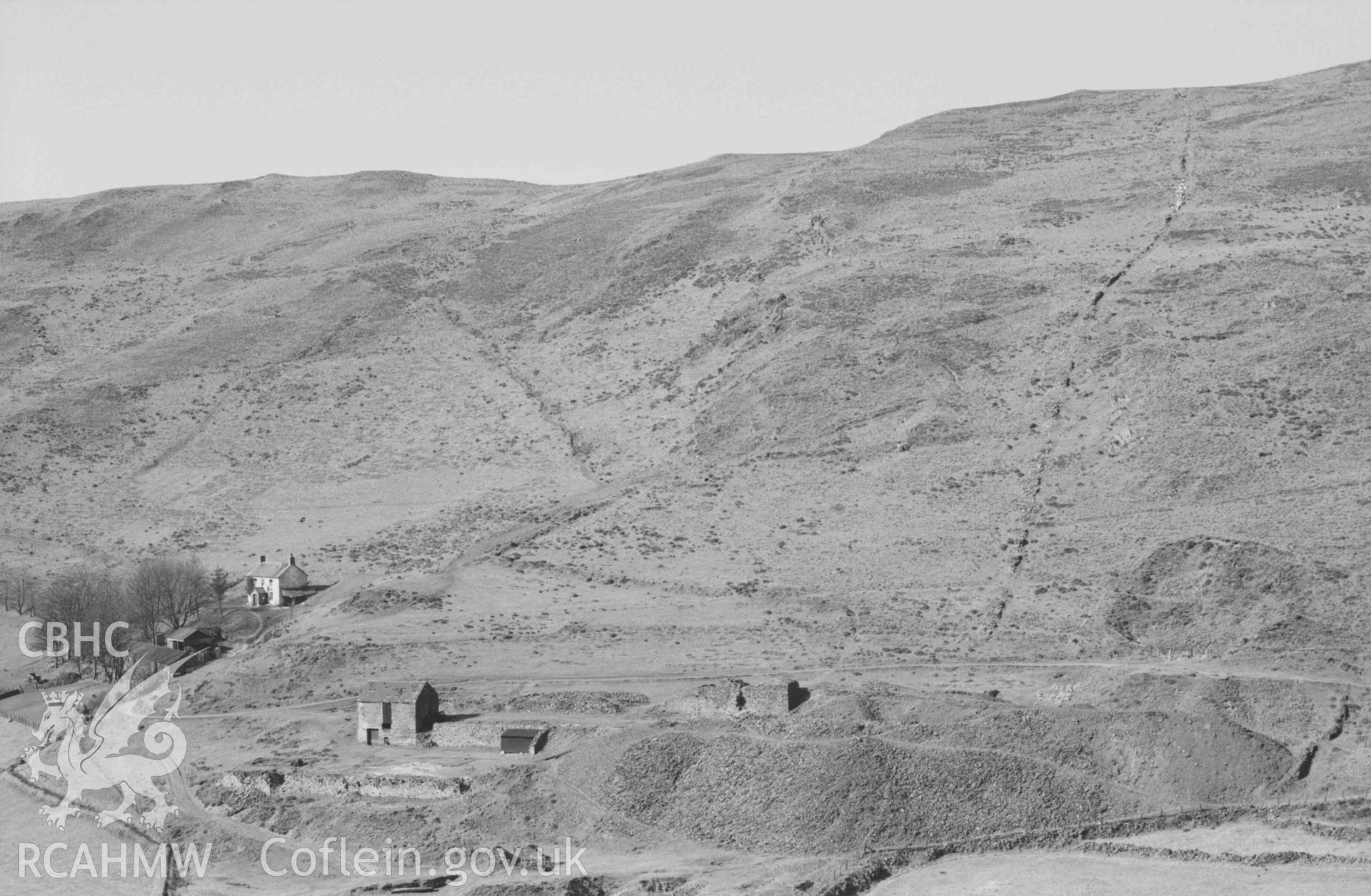 Digital copy of a black and white negative showing Llawr-y-cwm-bach farm and lead mine; very old, thick dry-stone wall on right running up Esgair Goch. Photographed by Arthur Chater on 6 April 1969. Looking north north west from Grid Reference SN 7080 8515.