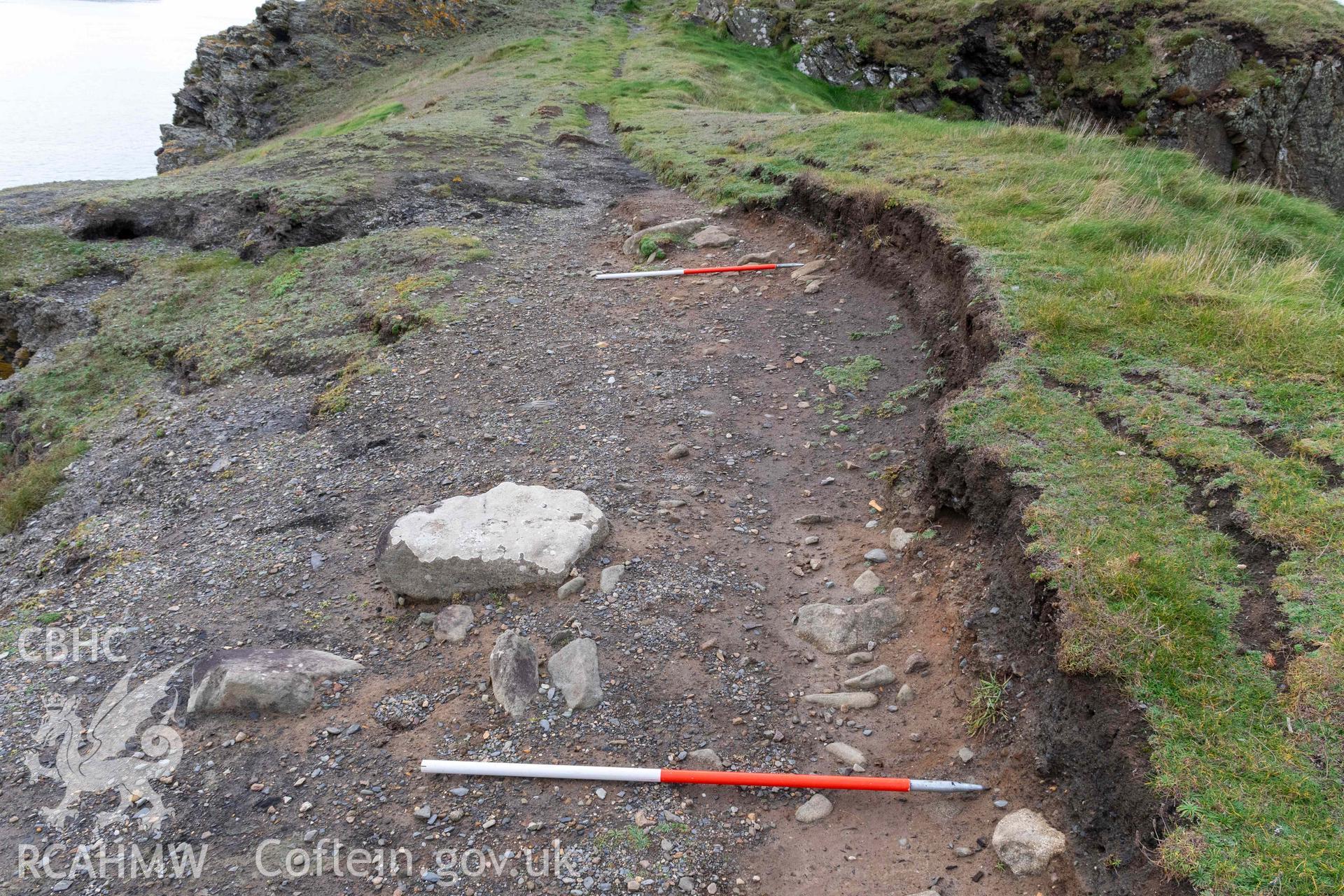 Trwyn Castell coastal promontory fort. View looking west across the narrowest point of the promontory where erosion has exposed walling of the forts defences (with scale).